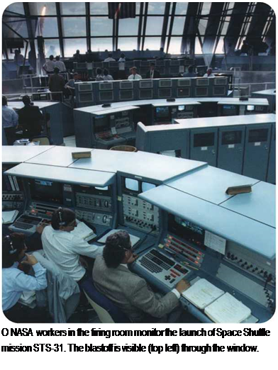 Подпись: O NASA workers in the firing room monitor the launch of Space Shuttle mission STS-31. The blastoff is visible (top left) through the window. 